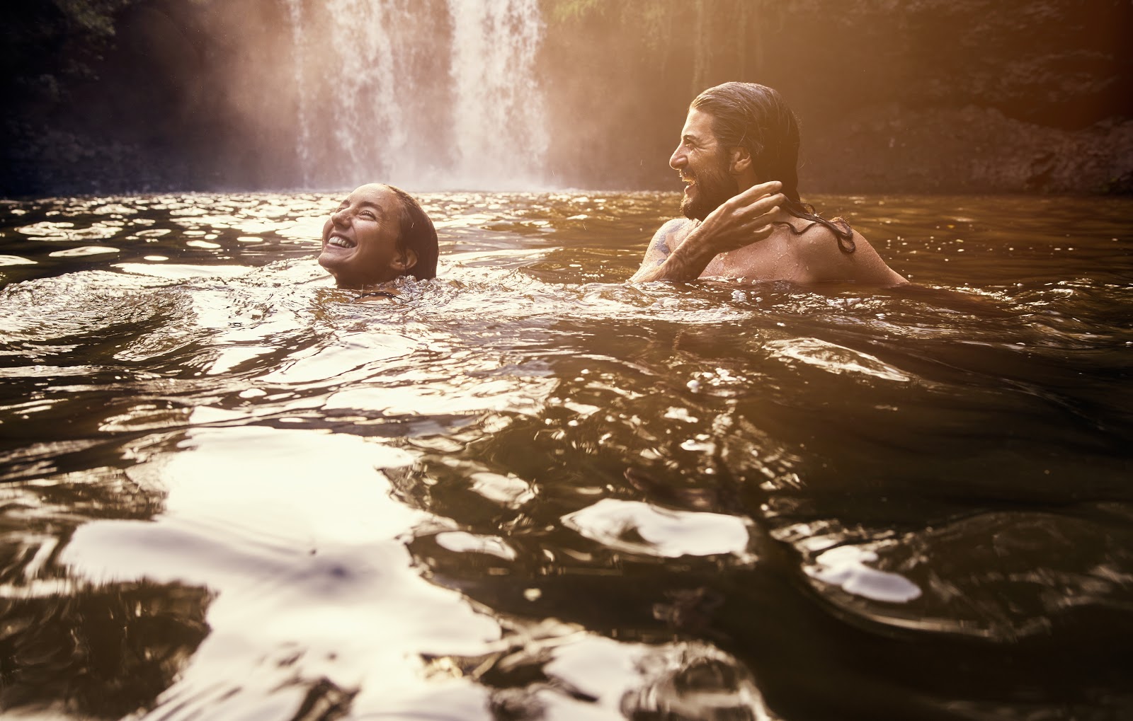 A man and woman smiling and swimming in a river by a waterfall.